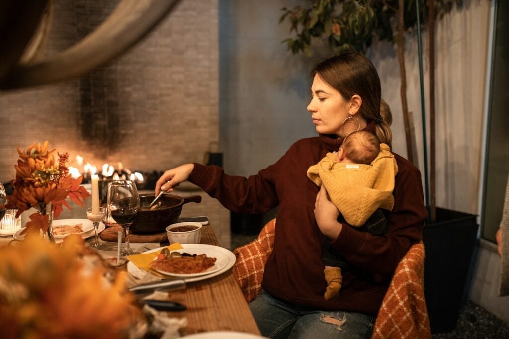 Woman in Maroon Long Sleeves Carrying a Baby While Eating